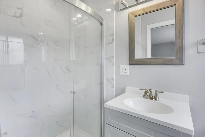 Glass-enclosed shower with white tiled walls, single vanity with integrated sink, wood-framed mirror, and brushed nickel fixtures