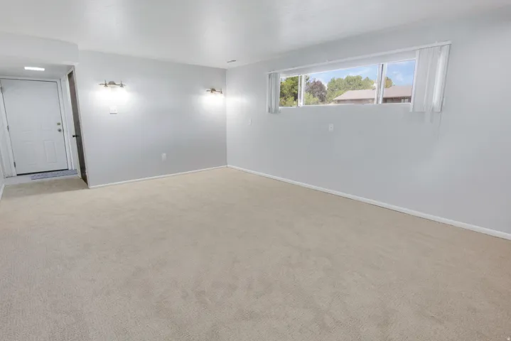 Expansive room featuring neutral carpeting, a white entry door, sconce lighting, and a window with blinds