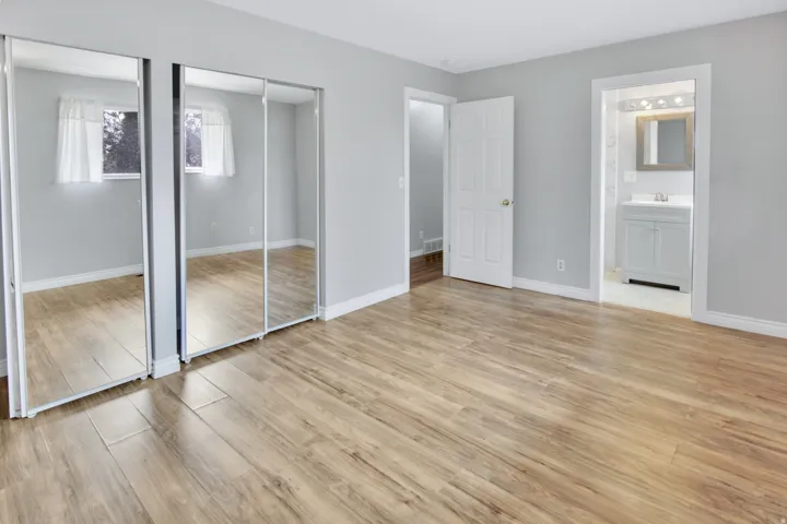 Wood-finish flooring throughout, light gray wall color, mirrored closet doors, white trim, and a full bathroom with a white vanity and framed mirror