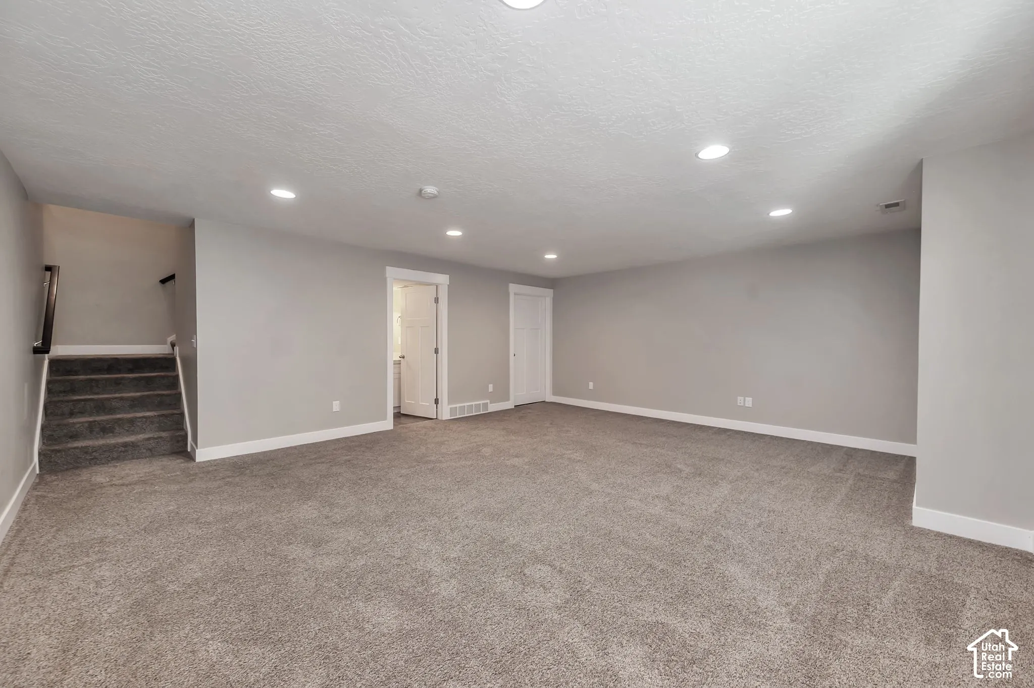 Spacious room featuring neutral carpeting, recessed lighting, and a staircase with a dark wood handrail