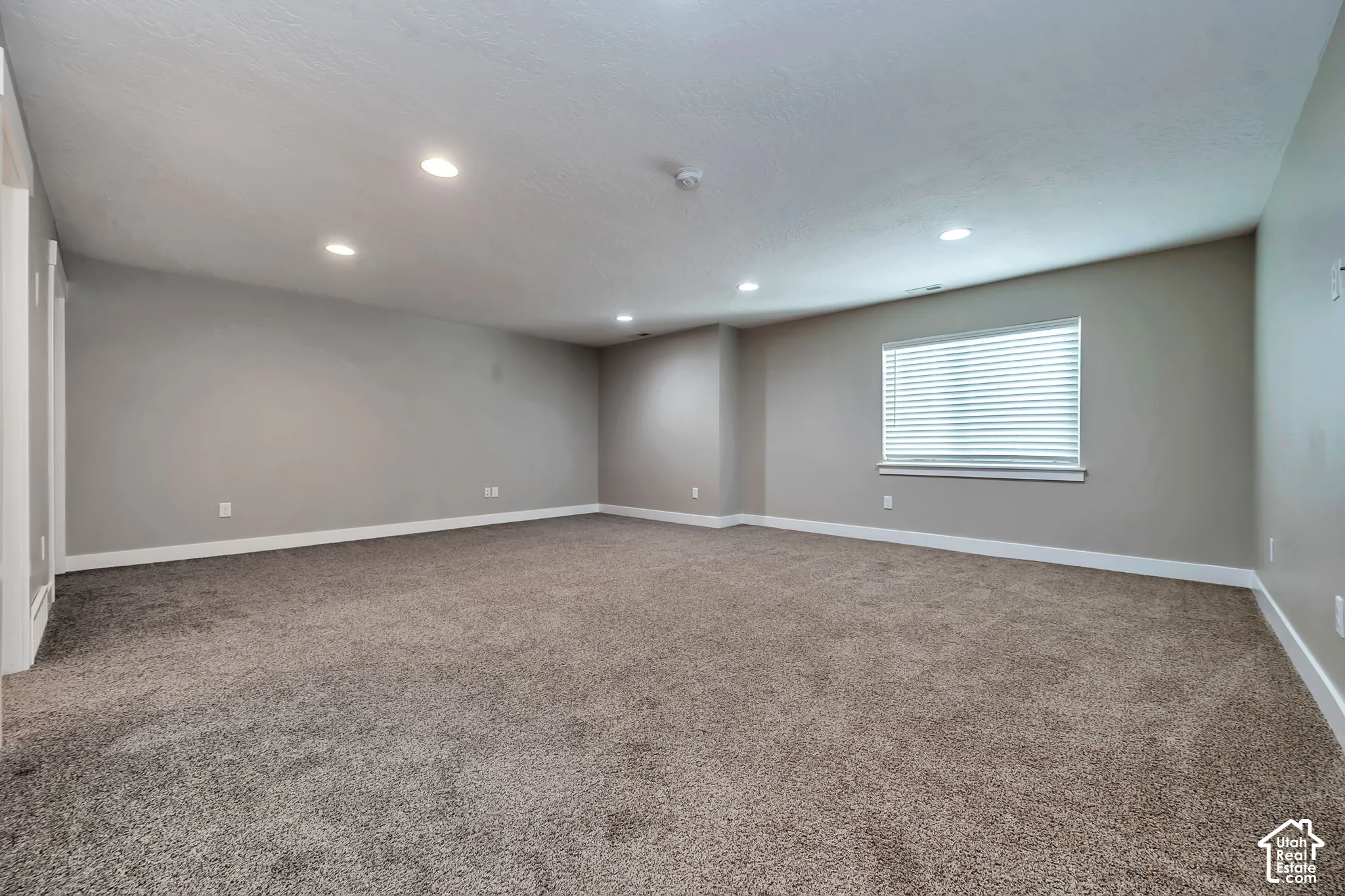 Spacious room with plush carpeting, recessed lighting, neutral wall tones, and a window with blinds