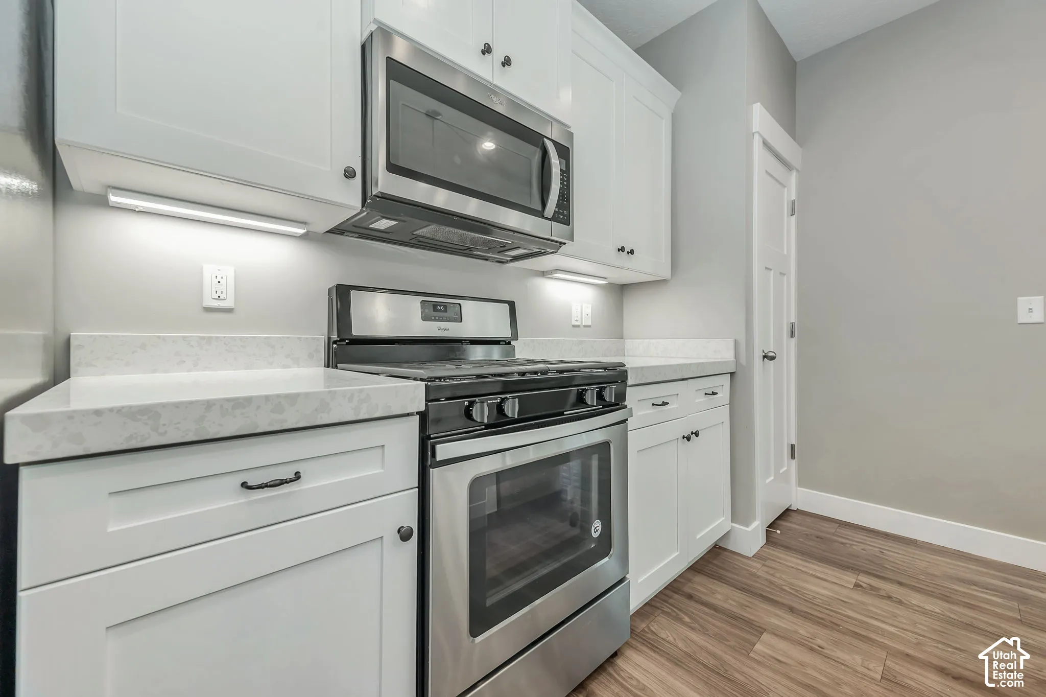 White shaker-style cabinetry with dark hardware, stainless steel range and microwave, light gray countertops, and wood-finish flooring