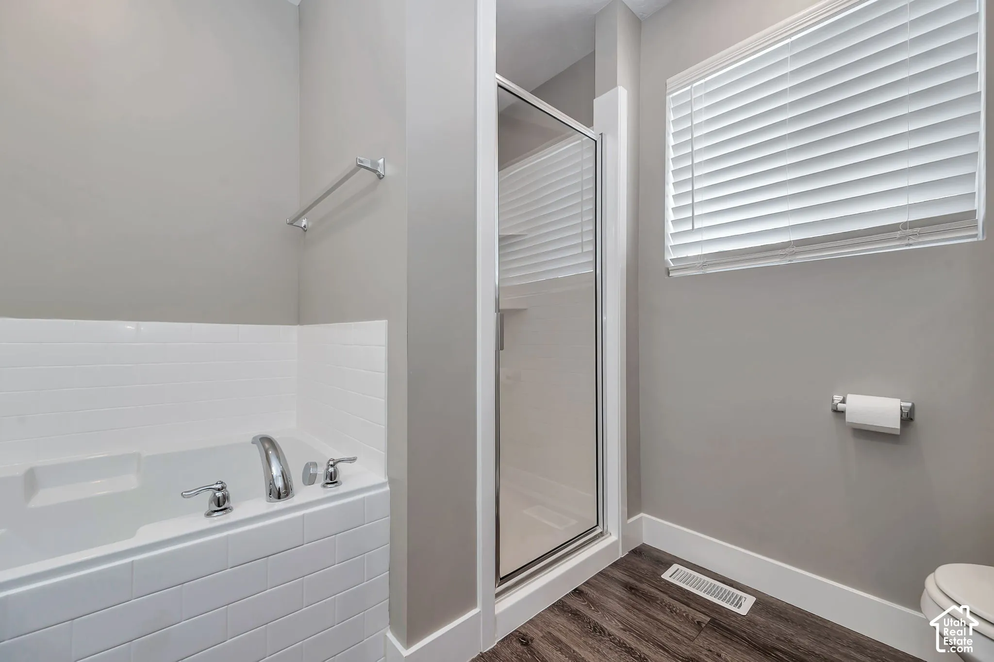 Built-in soaking tub with subway tile surround, separate glass-enclosed shower, wood-finish flooring, and window with blinds