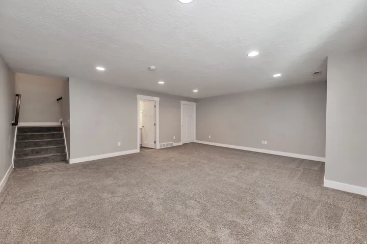 Spacious room featuring neutral carpeting, recessed lighting, and a staircase with a dark wood handrail