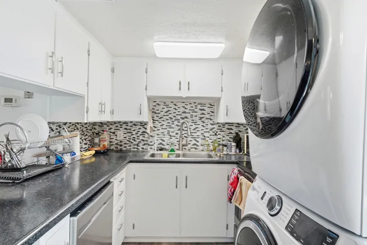 White cabinetry with brushed nickel hardware, black countertops, patterned tile backsplash, stainless steel dishwasher, and a stacked washer and dryer