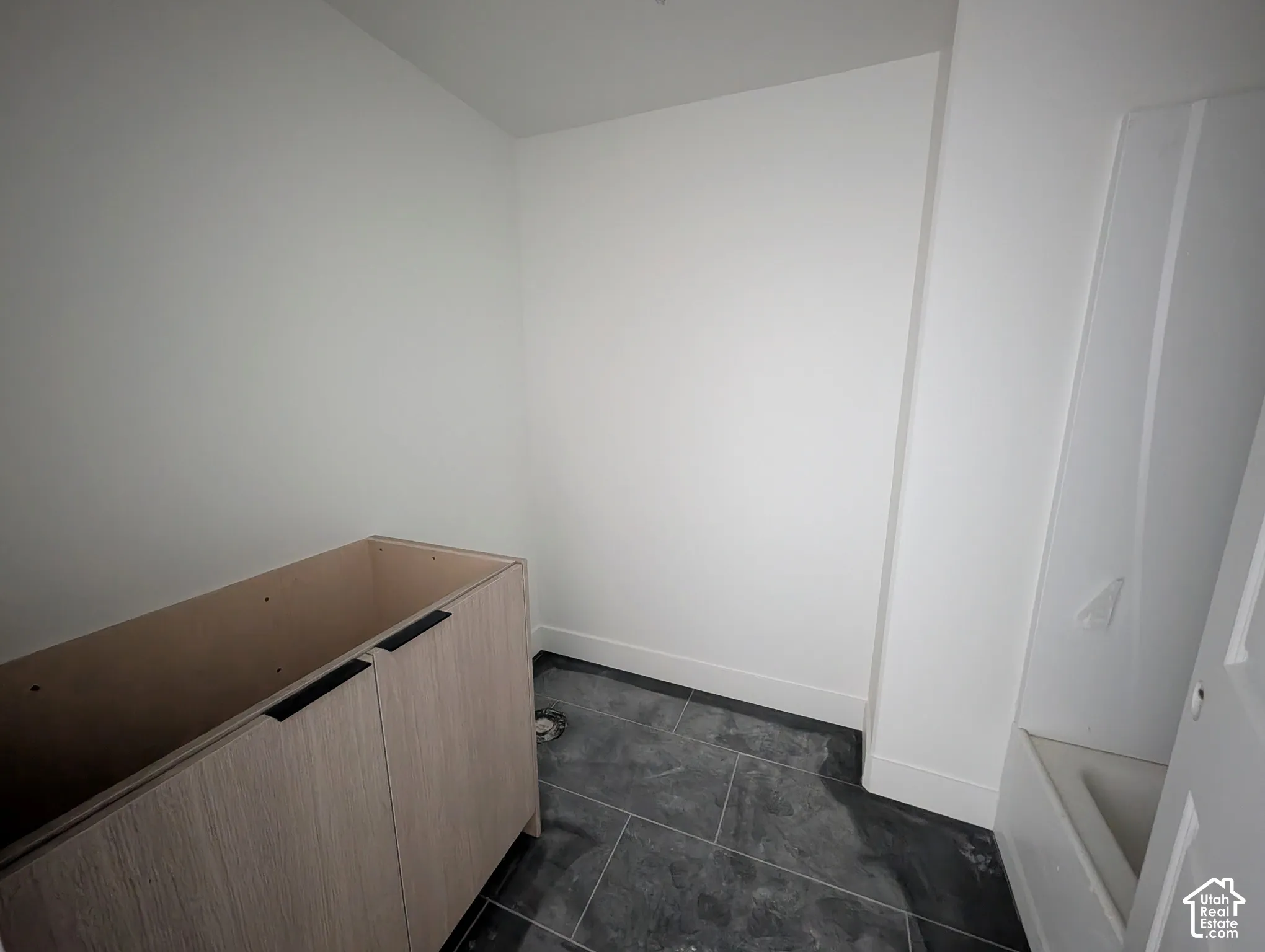 Unfinished vanity cabinet with two doors and matte black hardware, set against white walls and dark grey floor tiling