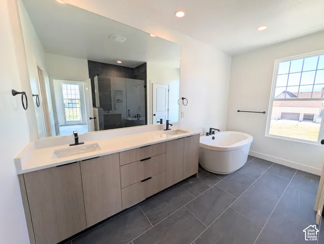 Spa-inspired primary bathroom featuring a dual-sink vanity with wood-finish cabinetry and white quartz countertops, frameless glass-enclosed shower with dark tile surround, freestanding soaking tub, slate-look tile flooring, and abundant natural light fro