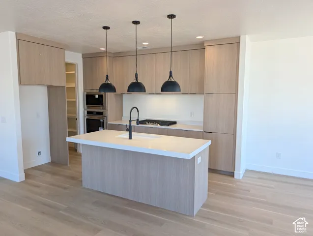 Contemporary kitchen featuring light wood-finish cabinetry, a central island with a solid surface countertop, built-in stainless steel double ovens, and sleek black pendant lighting