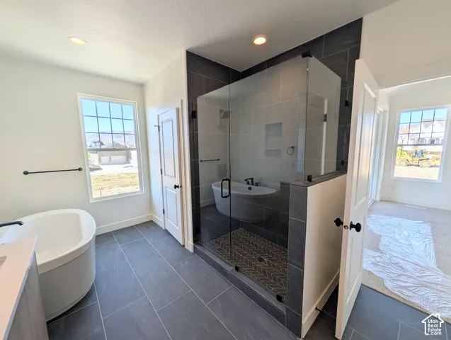 Freestanding soaking tub, walk-in shower with glass enclosure, dark gray tile flooring, recessed lighting, and white interior doors with black hardware