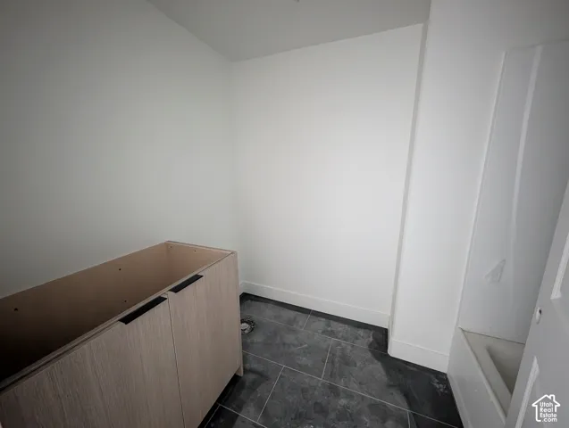 Unfinished vanity cabinet with two doors and matte black hardware, set against white walls and dark grey floor tiling