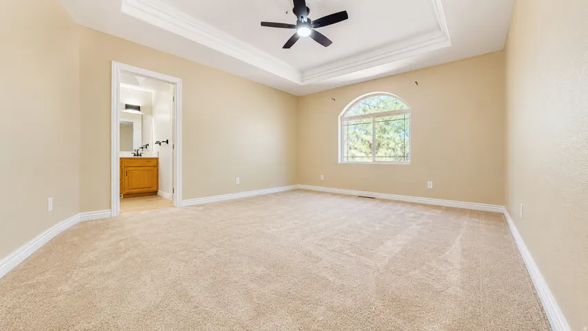 Expansive carpeted room featuring a tray ceiling with crown molding, ceiling fan, and arched window