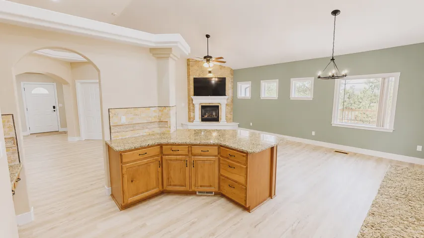 Kitchen island with light wood cabinetry and granite countertop, stone-faced fireplace, light wood-finish flooring, arched entryway, and multiple windows