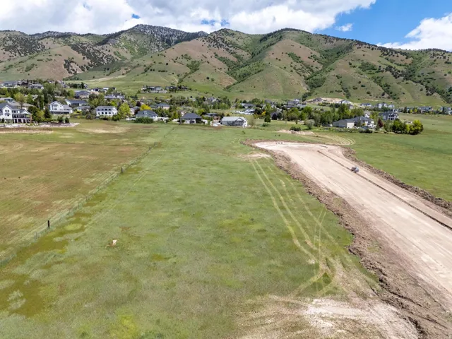 Expansive green field with a partial dirt road, surrounded by mountainous terrain and scattered residential properties