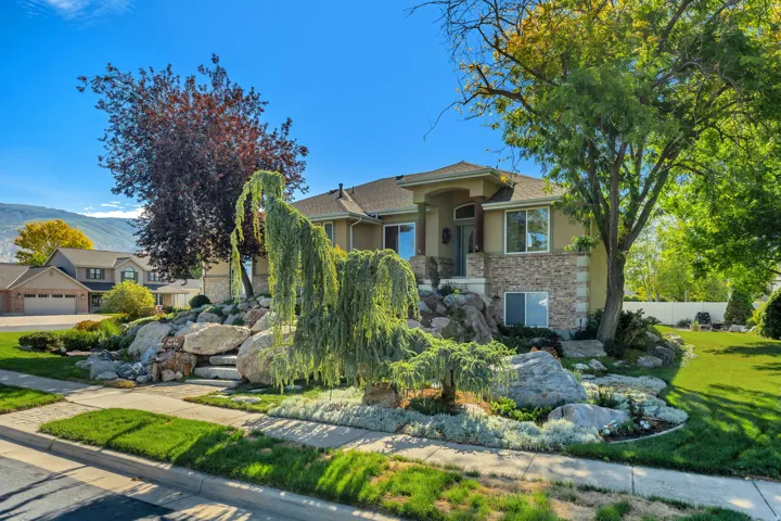 Stucco and stone facade home with a prominent entry, mature front yard landscaping, and a sloped terrain with large decorative boulders