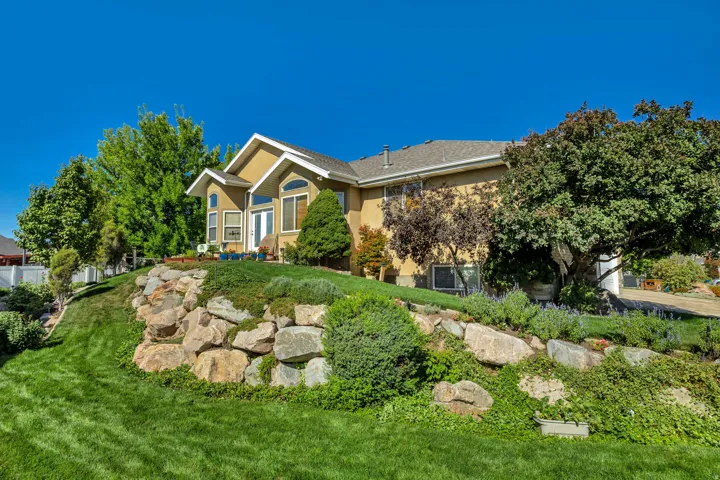 Stucco exterior with a pitched roof, prominent rock retaining wall, mature trees, and a manicured lawn