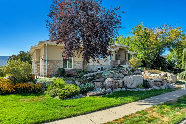Stucco and stone exterior with a covered entry, featuring a tiered rock garden, mature trees, and a concrete walkway