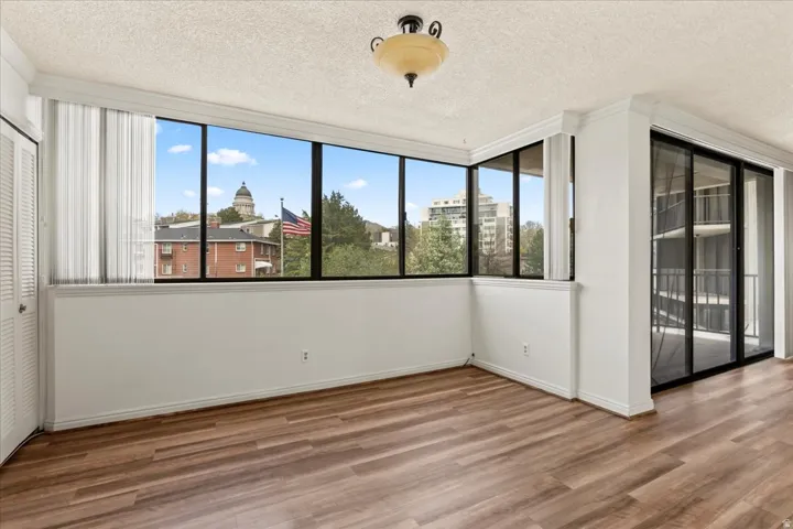 Expansive window wall with architectural backdrop, wide plank wood-finish flooring, and a sliding glass door to a private balcony