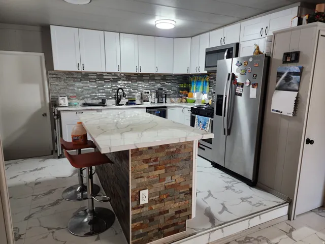 Kitchen featuring white shaker cabinetry, an iridescent tile backsplash, stainless steel appliances, a stone-clad island with a light-toned countertop, and patterned flooring