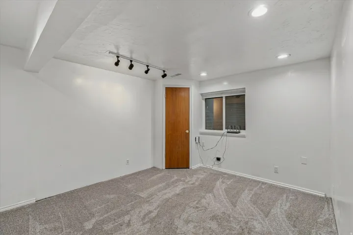 Bright, open room featuring recessed and track lighting throughout, neutral wall finishes, carpeted flooring, wood-finish entry door, and a window with integrated climate control unit