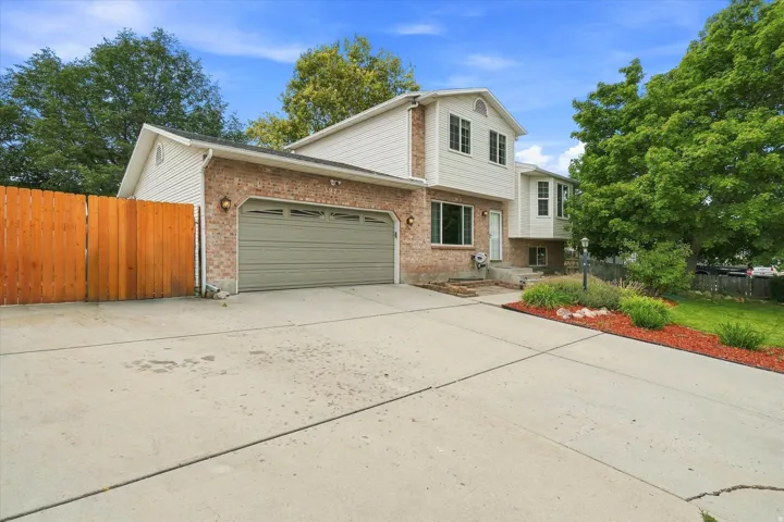 Two-story brick and siding home featuring a spacious concrete driveway, attached two-car garage with paneled door, and mature landscaping with established trees and manicured lawn areas bordered by decorative red mulch beds
