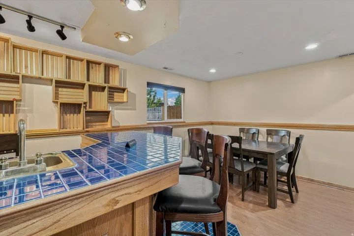 Finished basement recreation space featuring custom wood cabinetry with open shelving, blue ceramic tile flooring, wood-finish bar counter with seating, recessed and track lighting, and a wood dining table with chairs