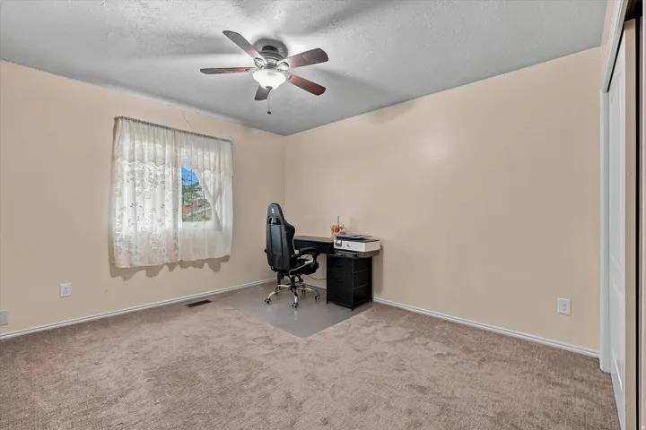 Spacious bedroom retreat featuring neutral wall finishes, plush carpeting, ceiling fan with integrated lighting, large window with natural light, and a dedicated workspace with built-in desk and storage cabinetry