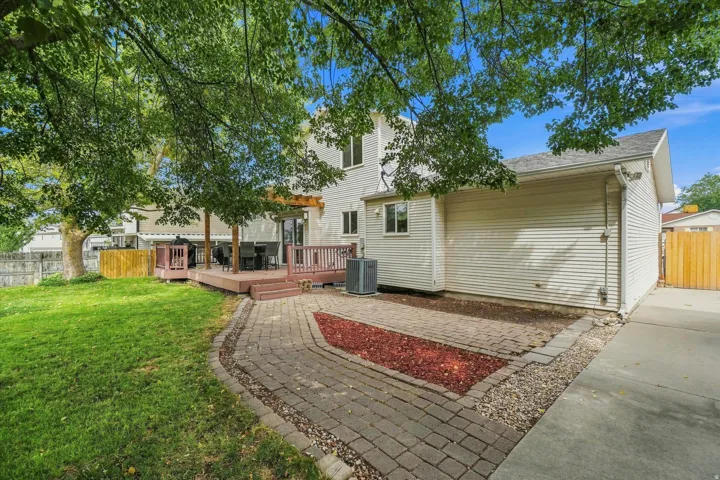 Spacious backyard oasis featuring mature shade trees, expansive manicured lawn, brick paver patio with integrated landscaping borders, wooden deck with railing, and detached garage