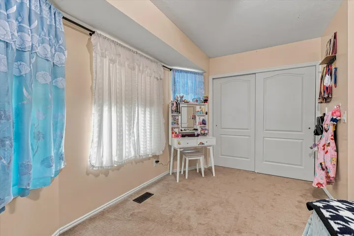 Spacious bedroom offering neutral wall finishes, carpeted flooring, large window with natural light, built-in closet storage with white panel doors, and a dedicated desk area with shelving