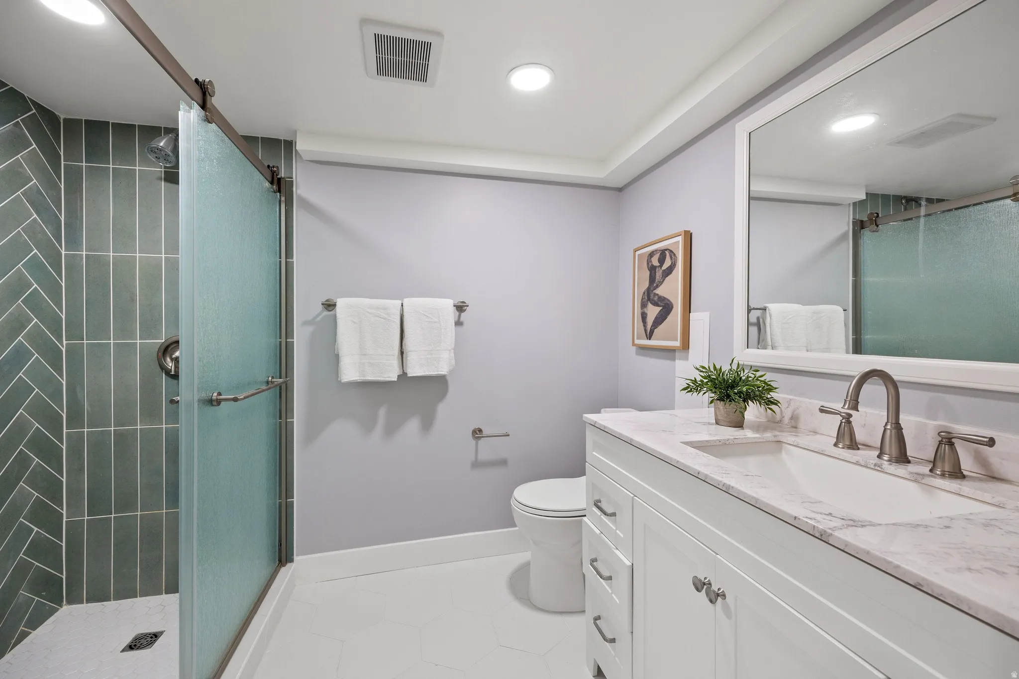 Shower with chevron tile and barn door style glass enclosure, white vanity with stone countertop, undermount sink, and brushed nickel fixtures