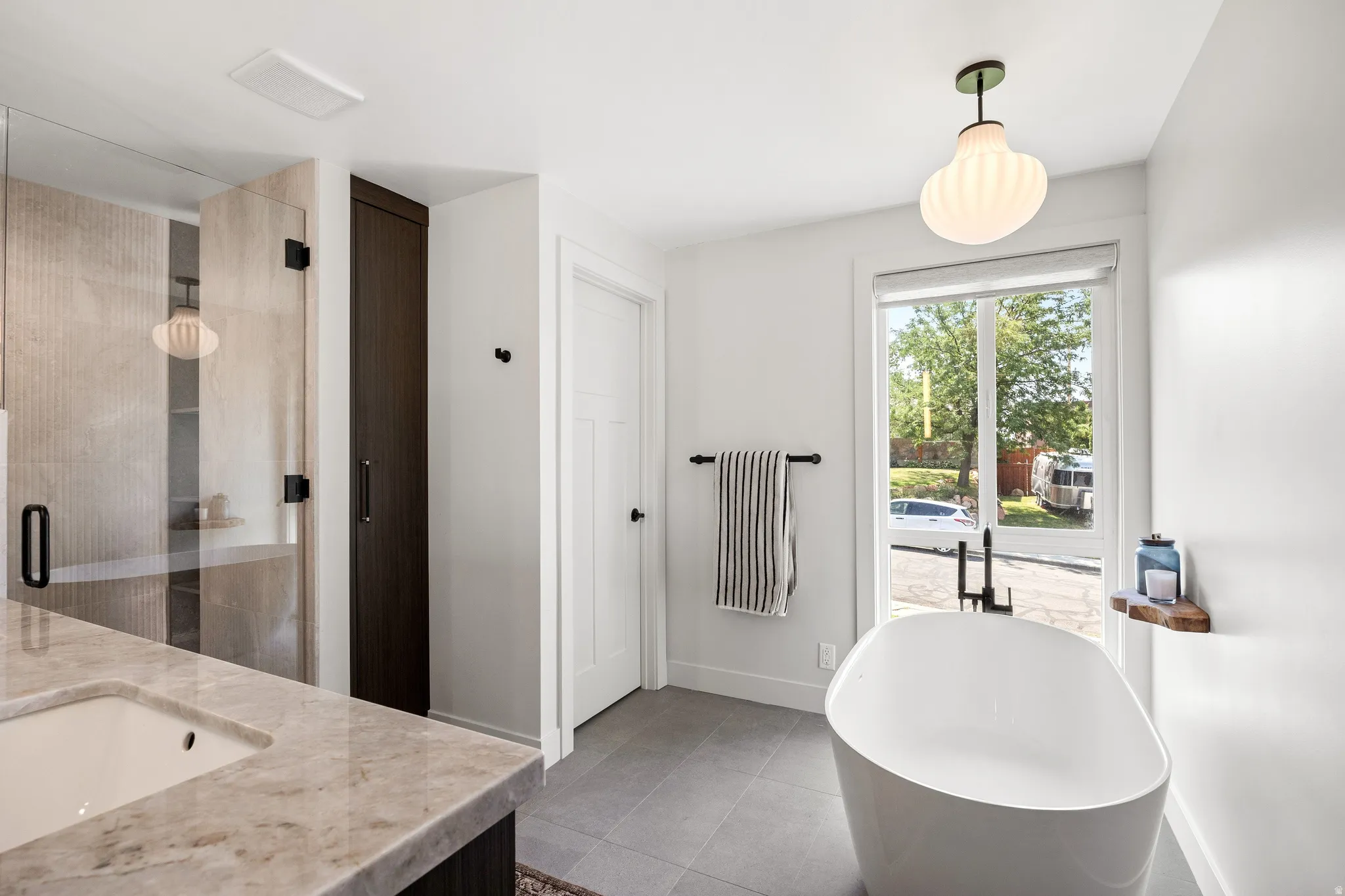 Freestanding soaking tub with a floor-mounted faucet, glass-enclosed shower, stone-finish vanity countertop, and dark wood-finish cabinetry