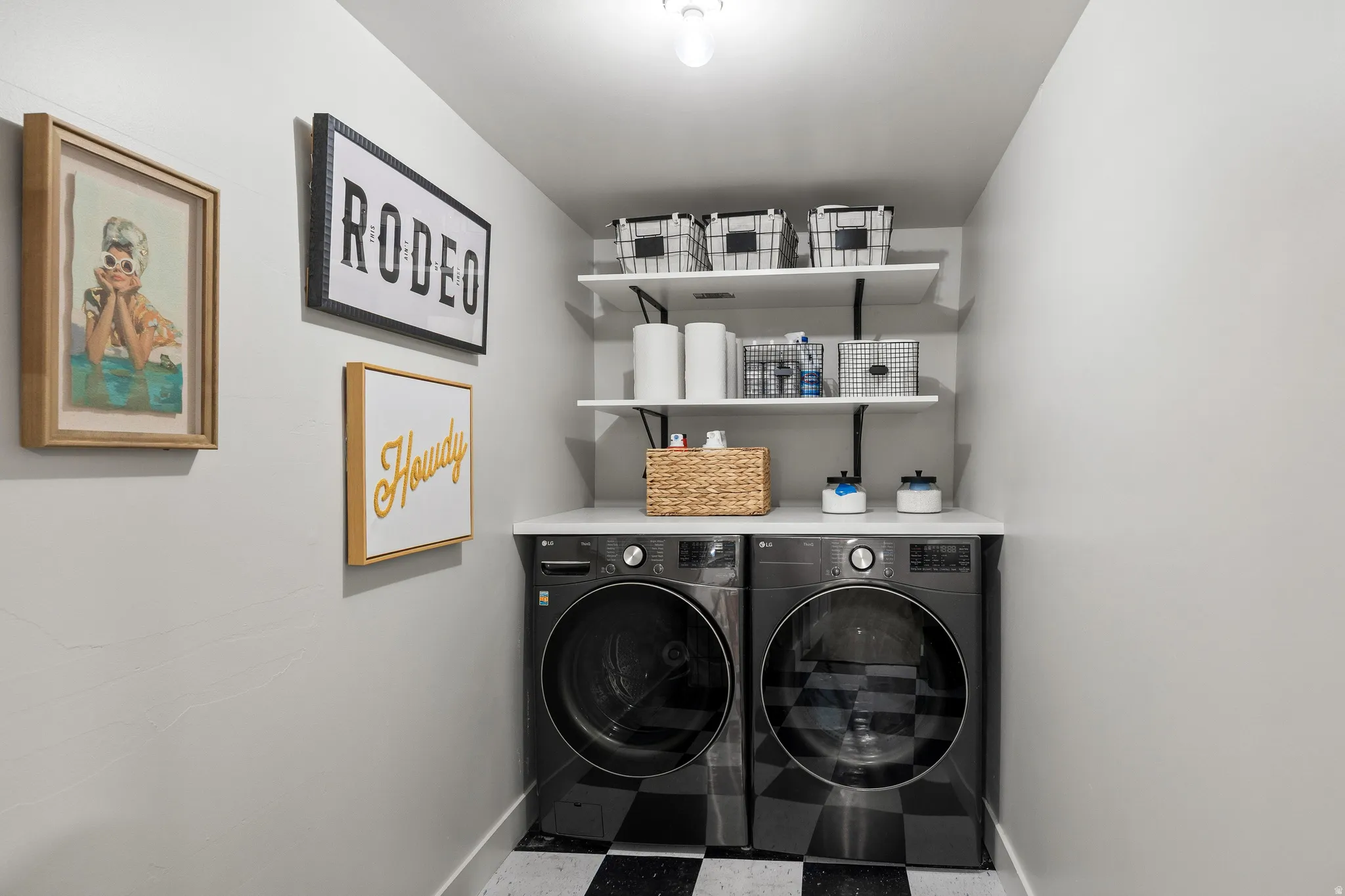 Dual laundry machines with a white countertop, wall-mounted shelving, and checkerboard flooring