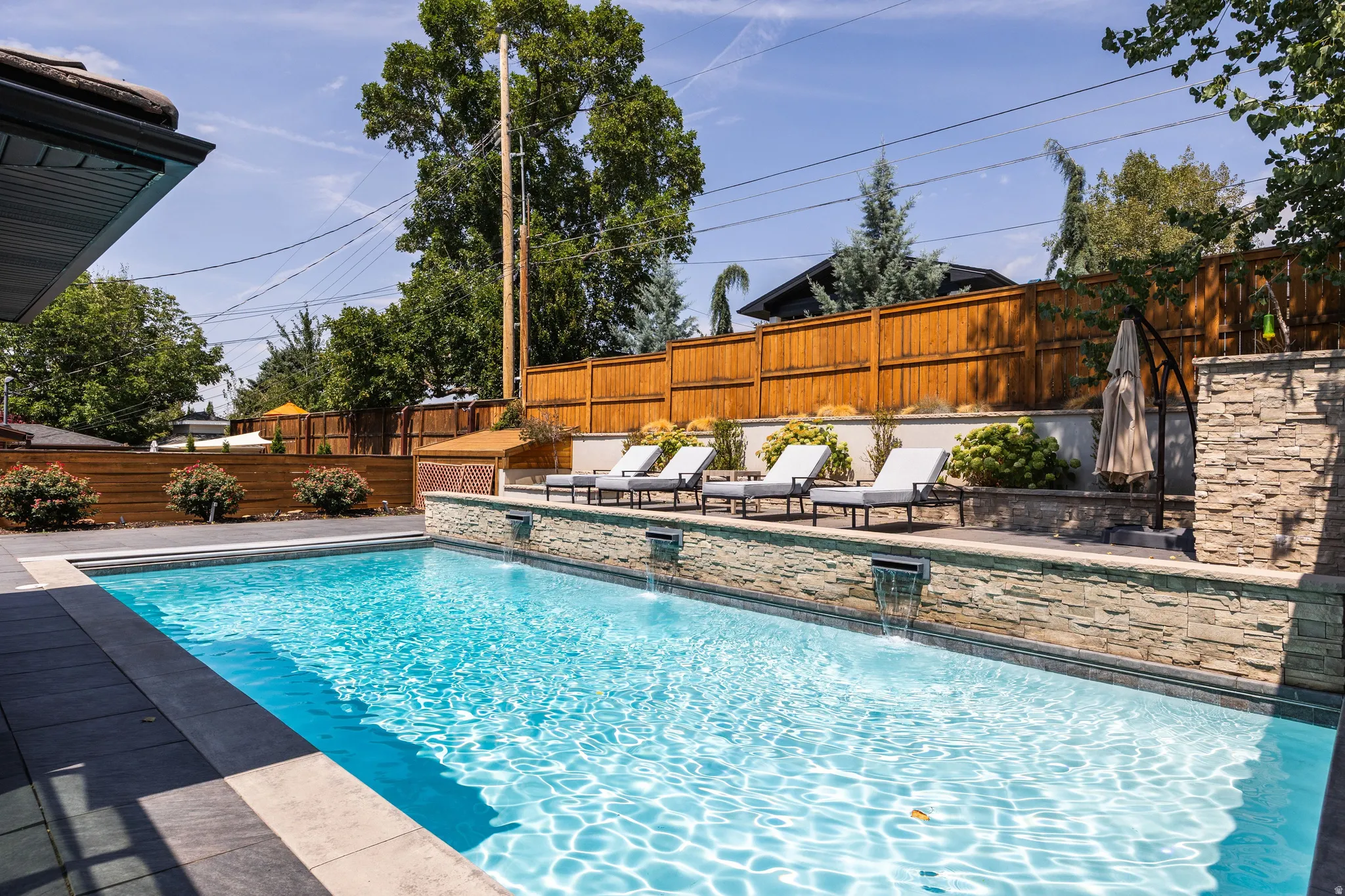 Outdoor swimming pool with built-in water features, stone-clad retaining wall, and wood fencing