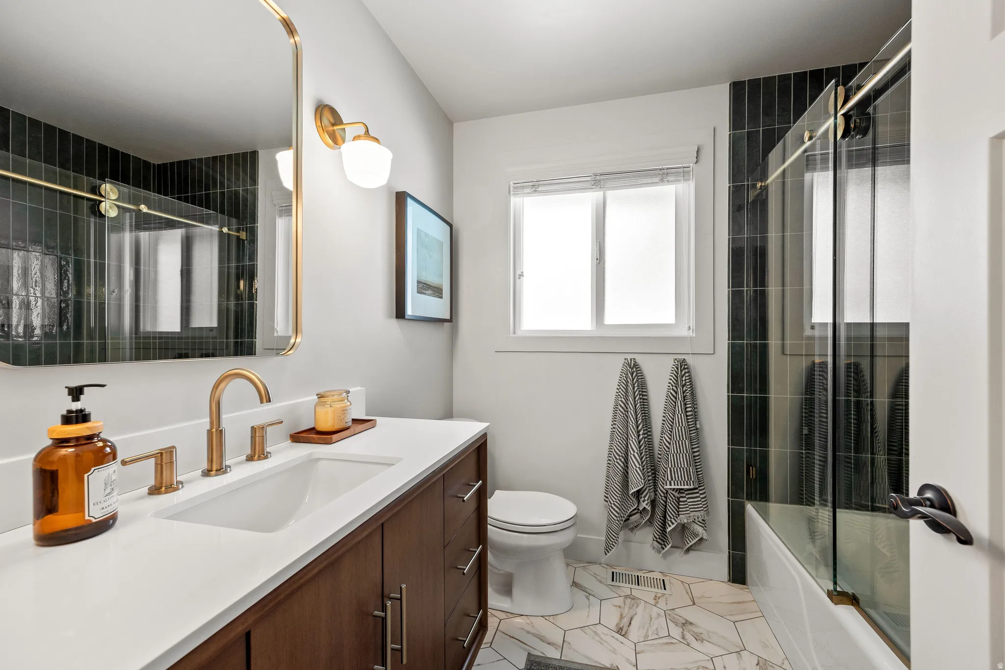 Dark green subway tile shower surround, brass-finish fixtures, white quartz-style vanity countertop, wood-finish vanity with drawer storage, and geometric tile flooring
