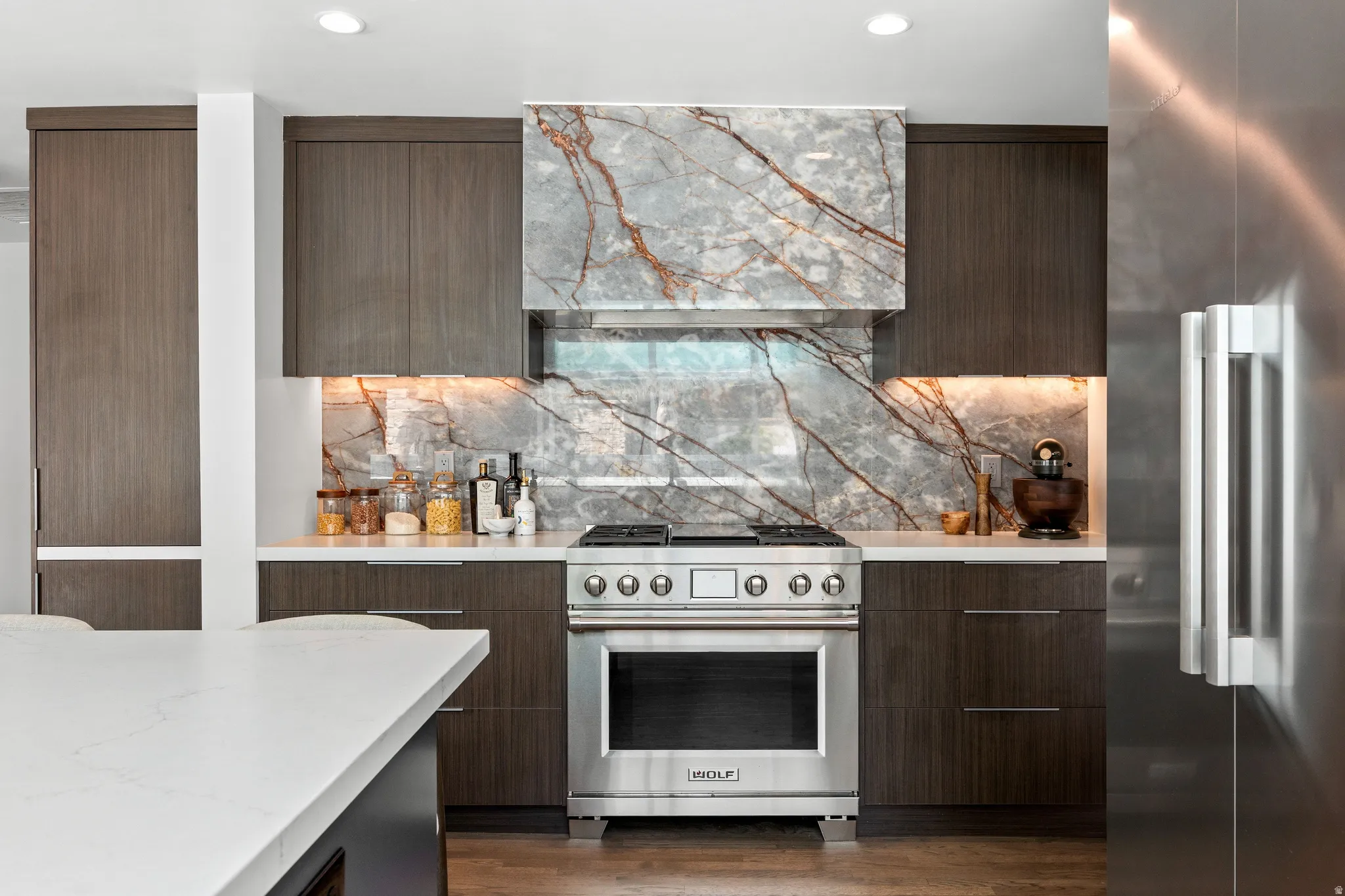 Stainless steel range with a custom stone hood, full-height stone backsplash, under-cabinet lighting, and wood-finish cabinetry