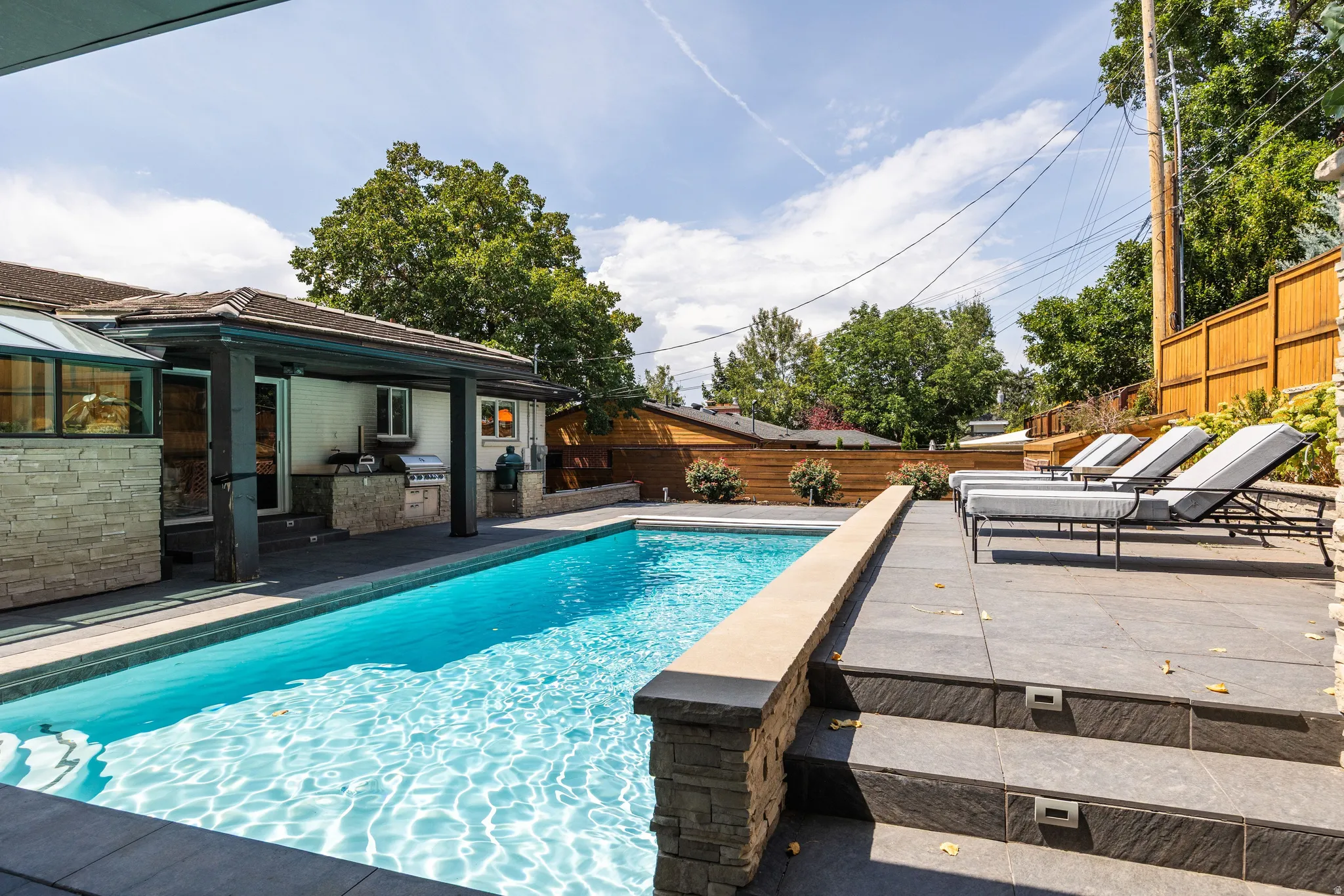 Outdoor swimming pool with tiled surround, stone accent wall, integrated outdoor kitchen, covered patio, and stepped hardscaping
