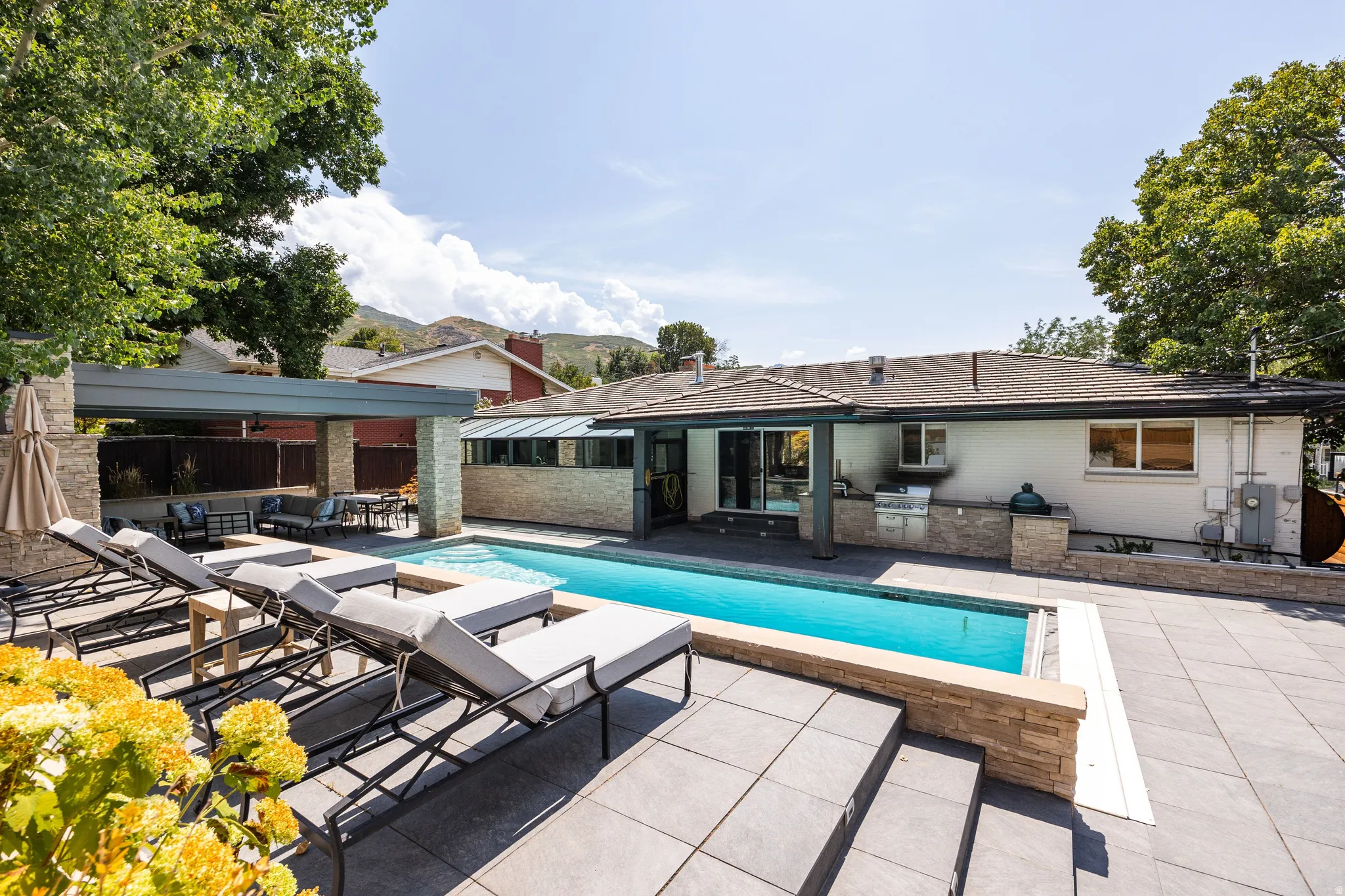 Backyard featuring a rectangular swimming pool, extensive stone paver patio, stone-clad outdoor kitchen, and a covered patio area with a built-in ceiling fan