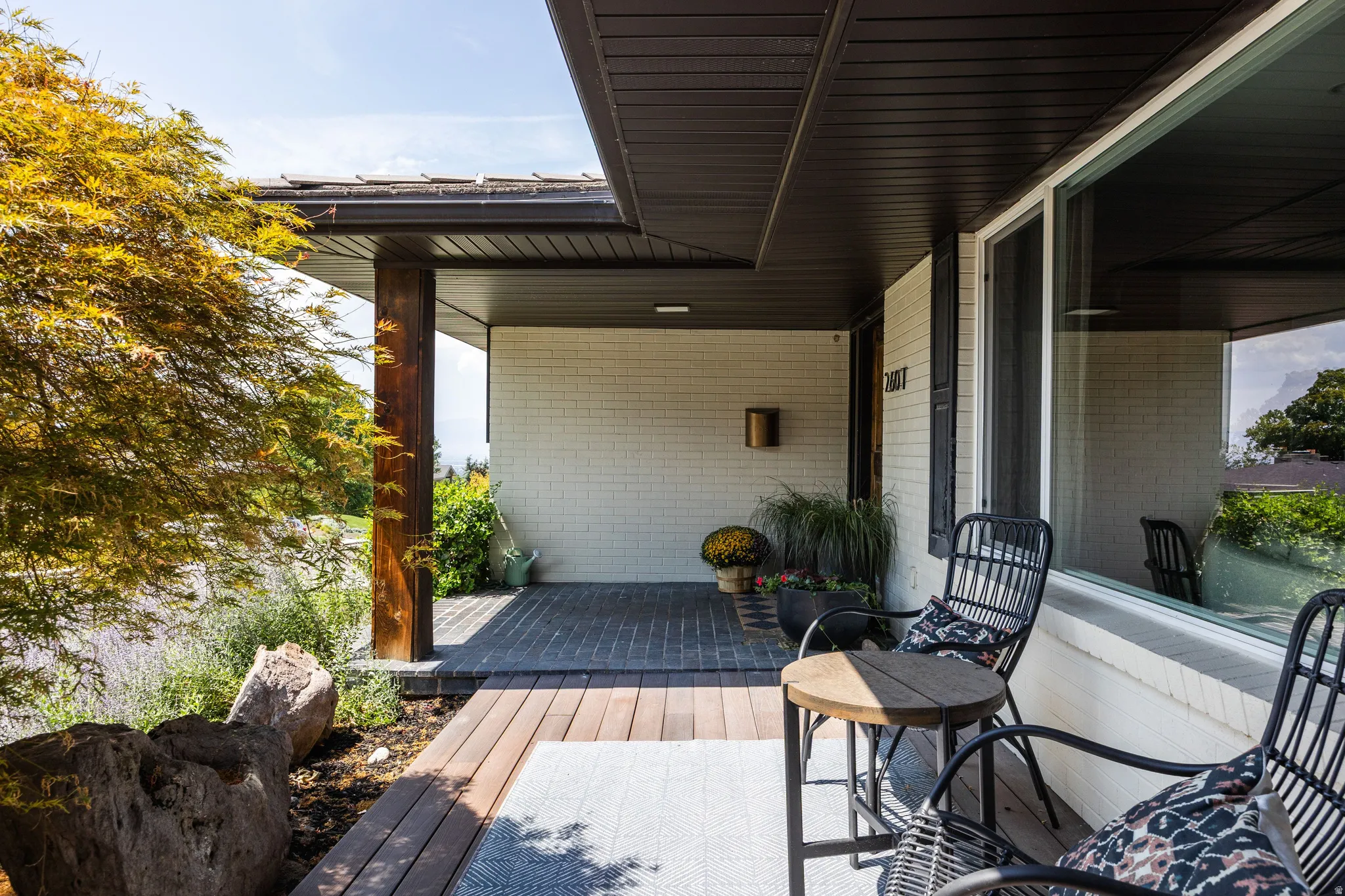 Covered front entry featuring a wood-plank patio, brick paving, dark soffits, and a large picture window
