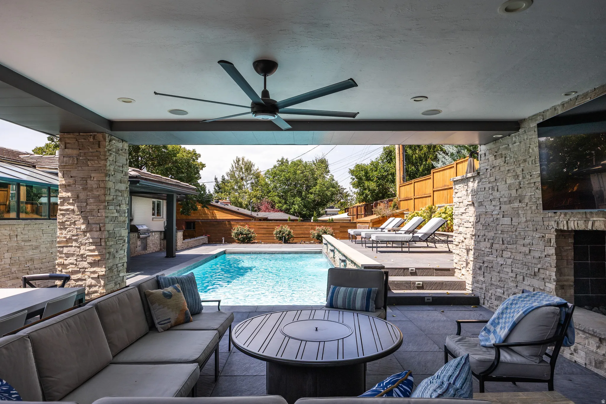 Covered patio with integrated lighting, a ceiling fan, stone-clad columns, a built-in outdoor fireplace, and a swimming pool