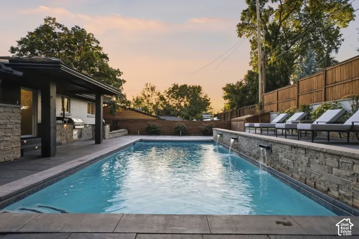 Rectangular in-ground swimming pool with water features, stone retaining wall, and an outdoor kitchen area