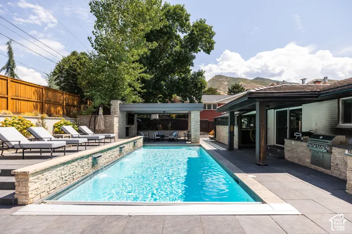 Rectangular swimming pool with water features, stone-clad pool surround, covered outdoor living space, and an integrated outdoor kitchen with a built-in grill