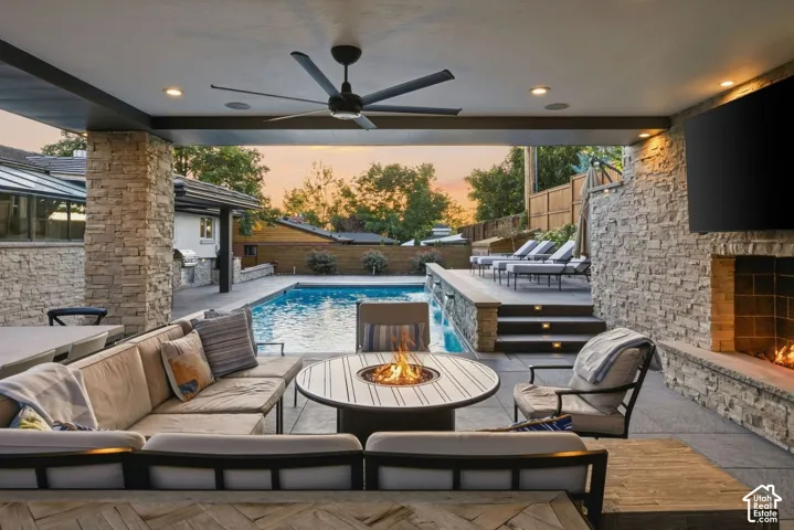 Covered outdoor living space with integrated ceiling fan, stacked stone fireplace, built-in outdoor kitchen, swimming pool, and tiered hardscaping