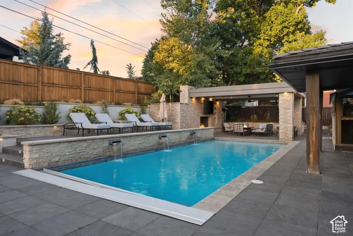 Rectangular swimming pool with stone waterfall features, integrated stone patio with a covered seating area, built-in outdoor fireplace, and dark-toned paver decking