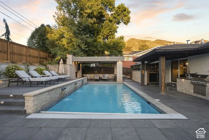 Outdoor swimming pool with water features, covered patio, built-in outdoor kitchen with grill, and stone-clad retaining walls