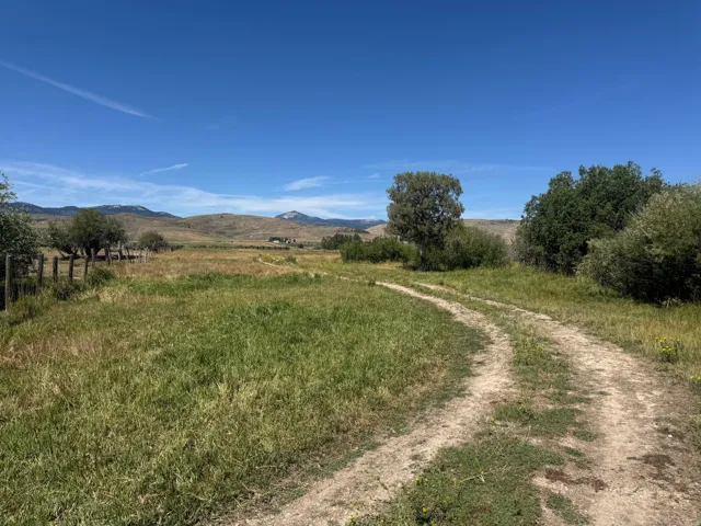 Expansive open land with a dirt path, featuring distant mountainous terrain, scattered trees, and a wooden fence line