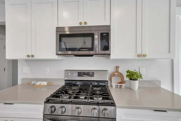 White shaker-style cabinetry with brass hardware, stainless steel gas range, coordinating stainless steel microwave, and light-toned countertops