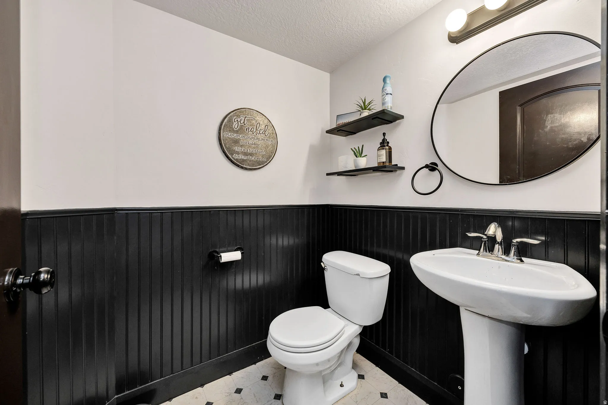 Elegant powder room featuring black beadboard wainscoting, a pedestal sink with chrome fixtures, a low-profile toilet with closed lid, and a large round mirror with metal frame
