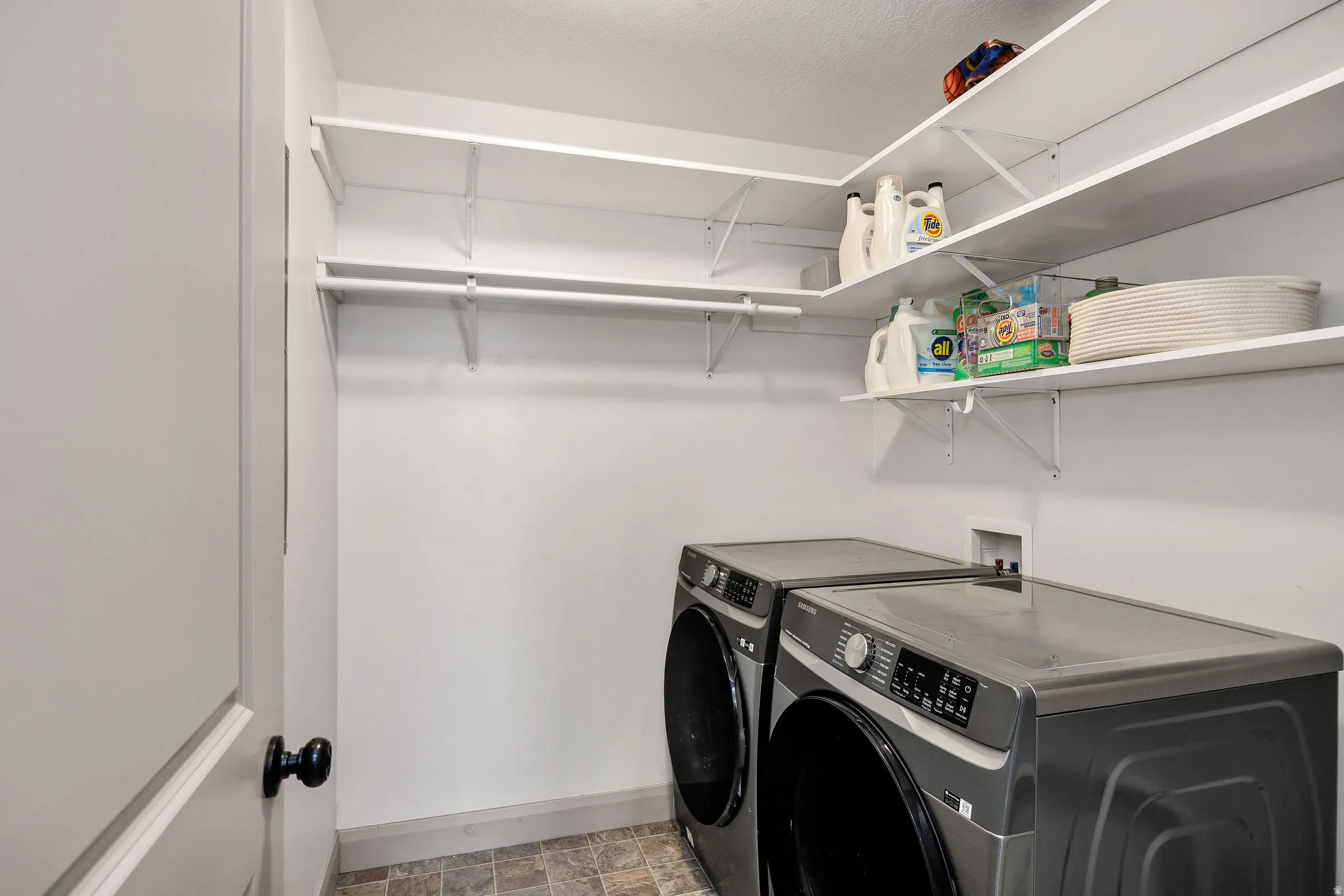 Dedicated laundry room featuring stacked front-load appliances with digital controls, ceramic tile flooring, white painted walls, and ample open shelving with metal brackets for storage and organization