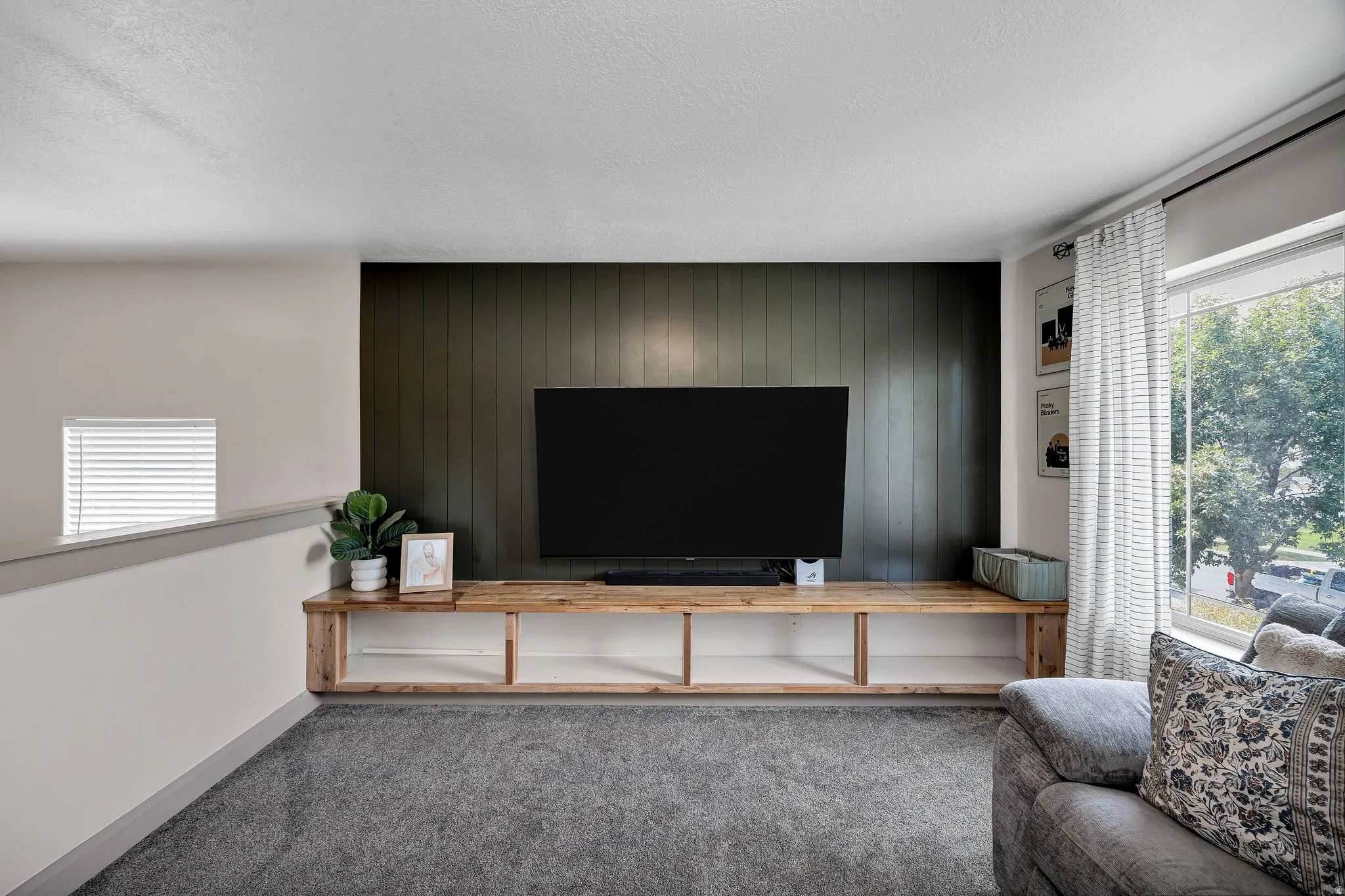 Built-in media console with open shelving, dark vertical panel accent wall, large window with roller shades, and plush gray carpeting
