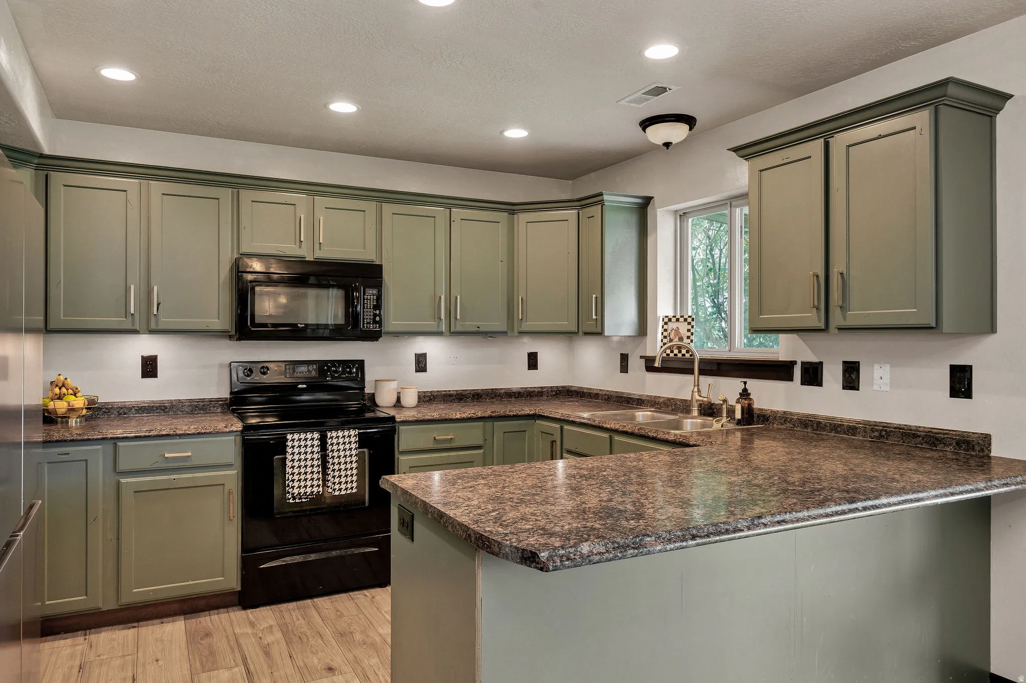 Chef's kitchen featuring sage-green cabinetry with brushed nickel hardware, granite countertops, a substantial island with seating space, stainless steel appliances including a black range and microwave, recessed lighting, and wood-finish flooring through