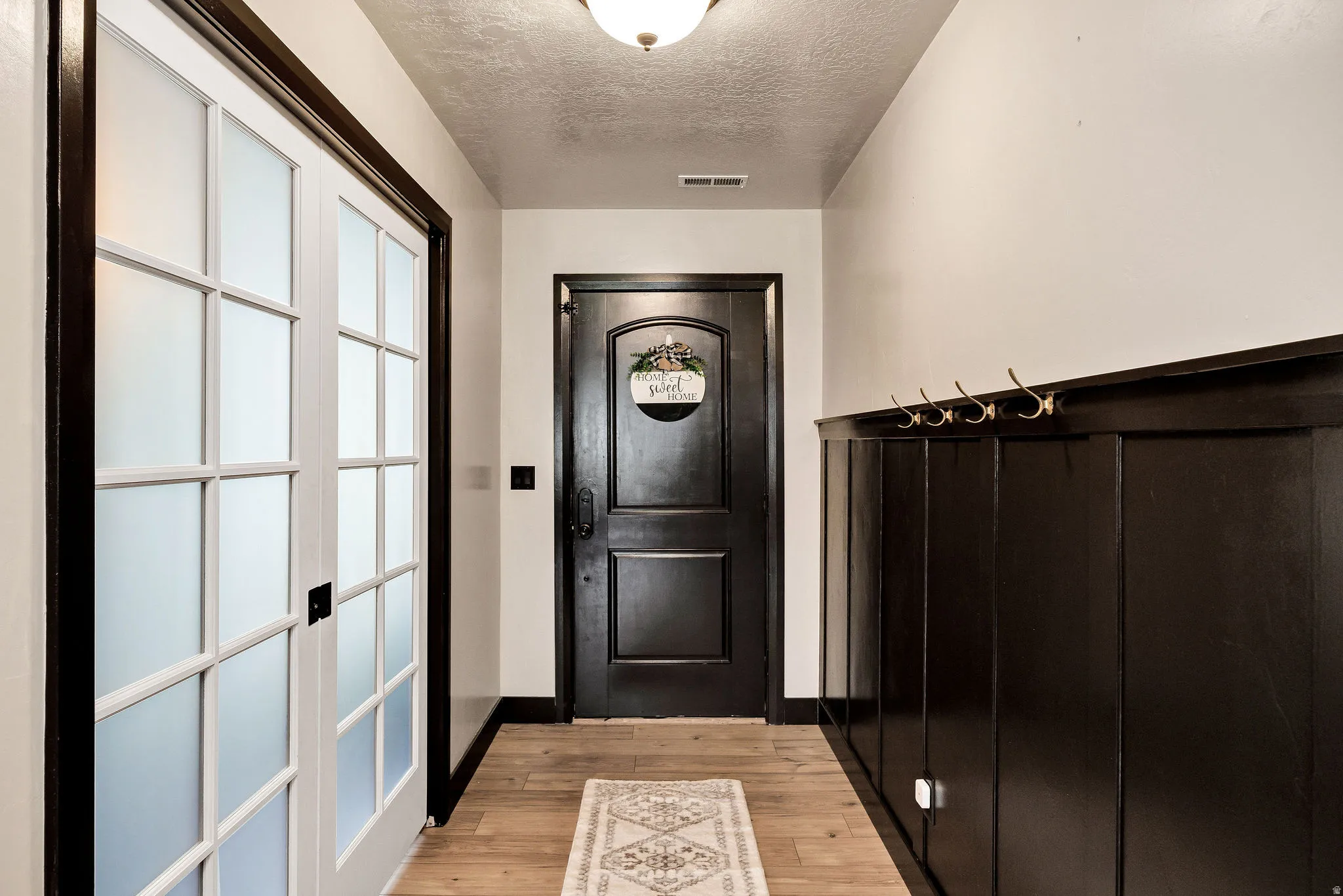 Wood-finish flooring, white French doors with frosted glass, dark wood paneling, dark arched entry door, and gold-tone wall hooks